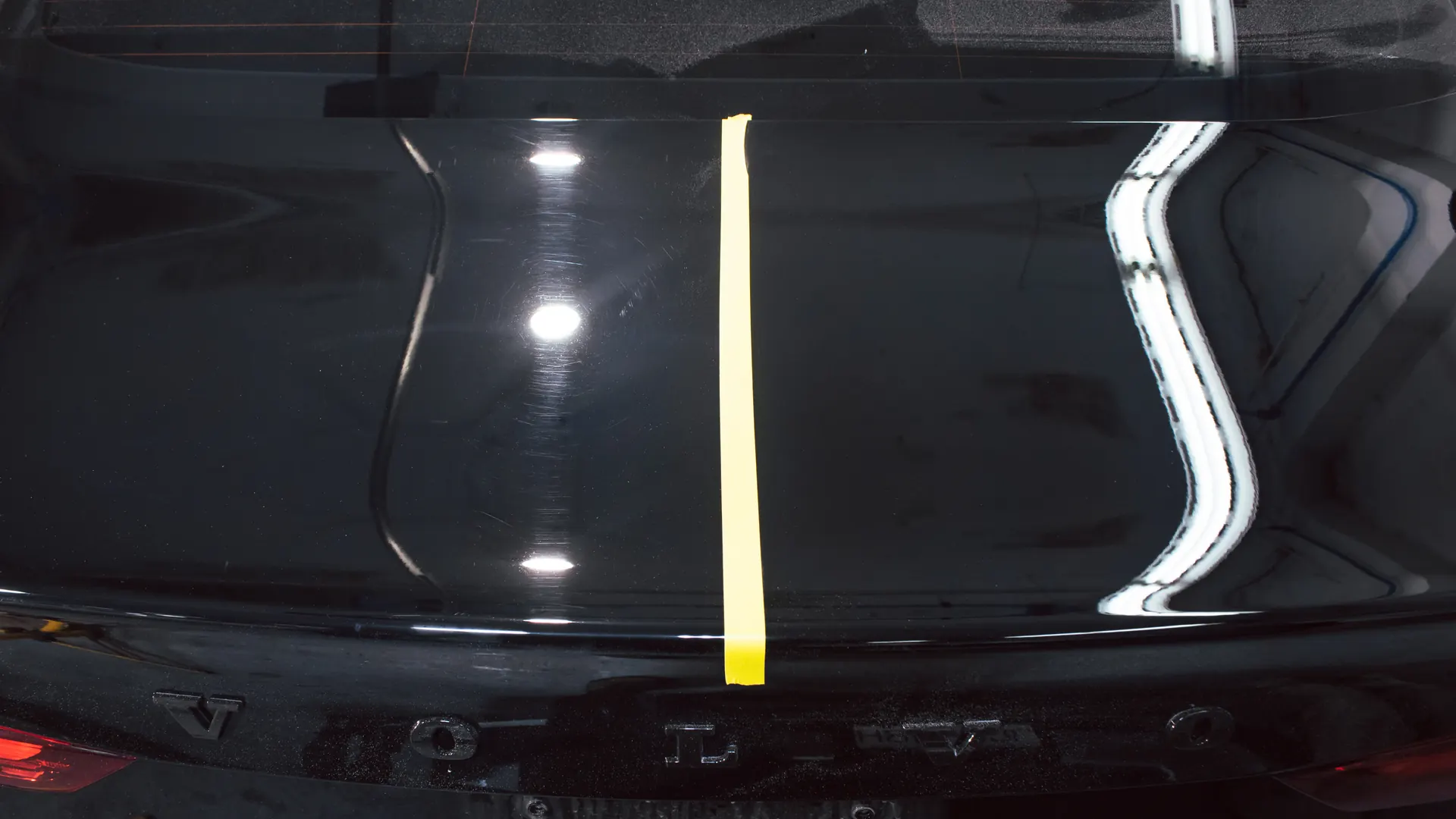 Black Volvo tailgate divided by tape to show paint swirls before correction and improved clarity after correction